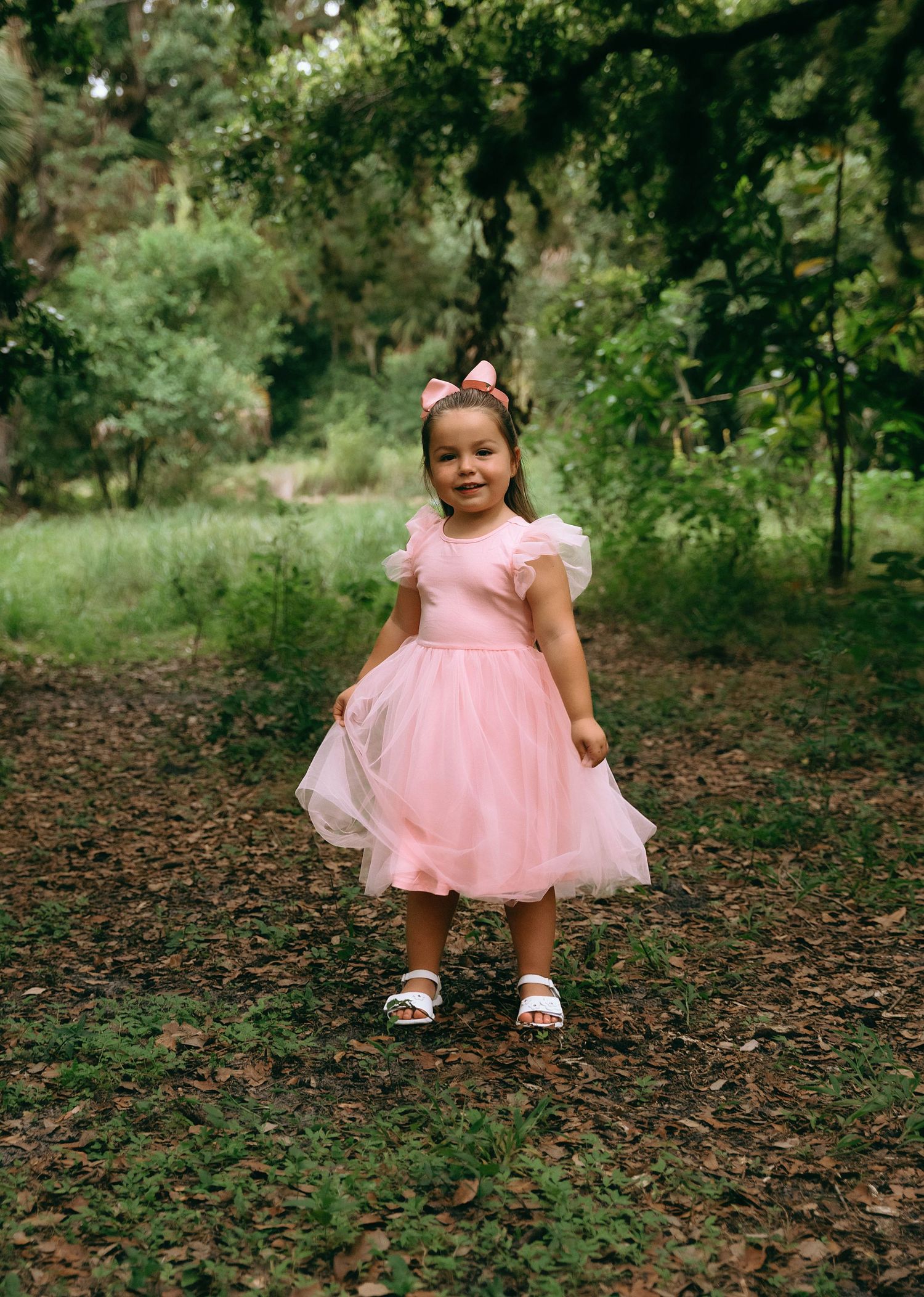 A pink tulle dress creates a whimsical scene against fallen leaves in a wooded outdoor setting.