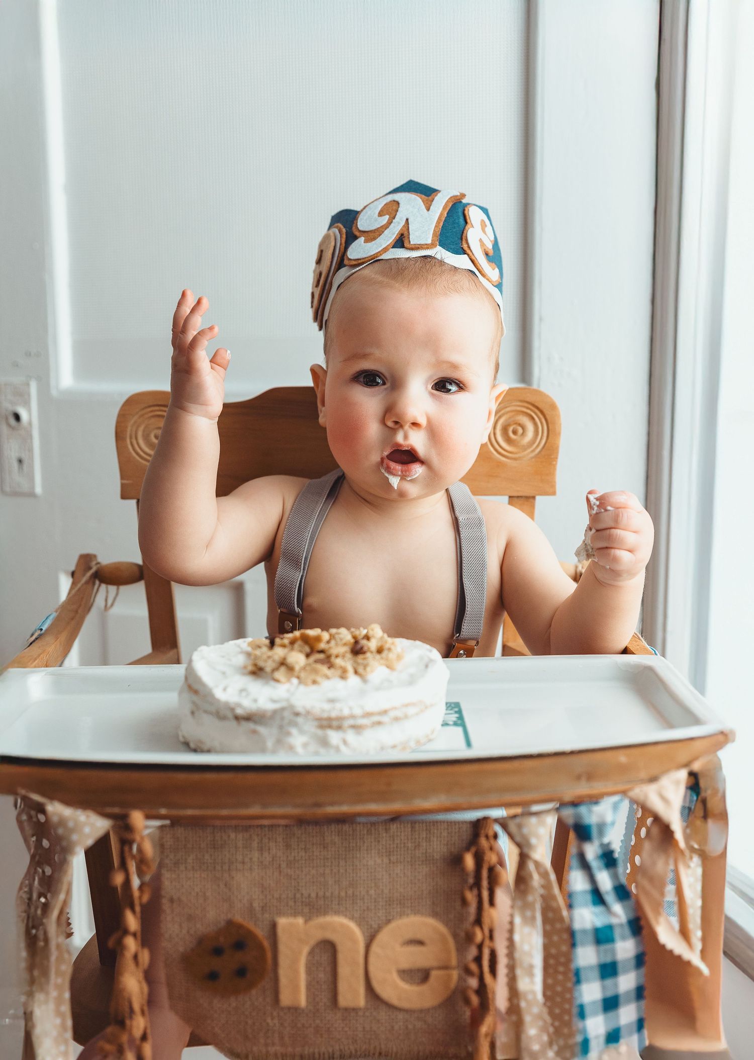 A baby wearing a patterned hat sits in a vintage wooden high chair ready to eat their first birthday cake.