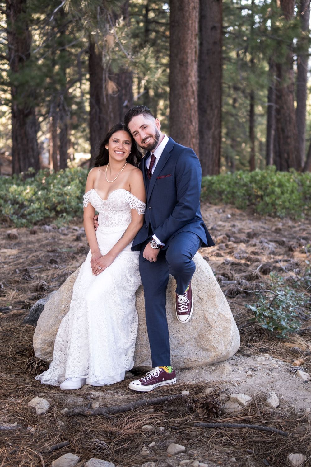 A wedding couple sits on a large boulder in a forest setting by Tannenbaum Reno Event Center, the groom wearing sneakers with his suit.