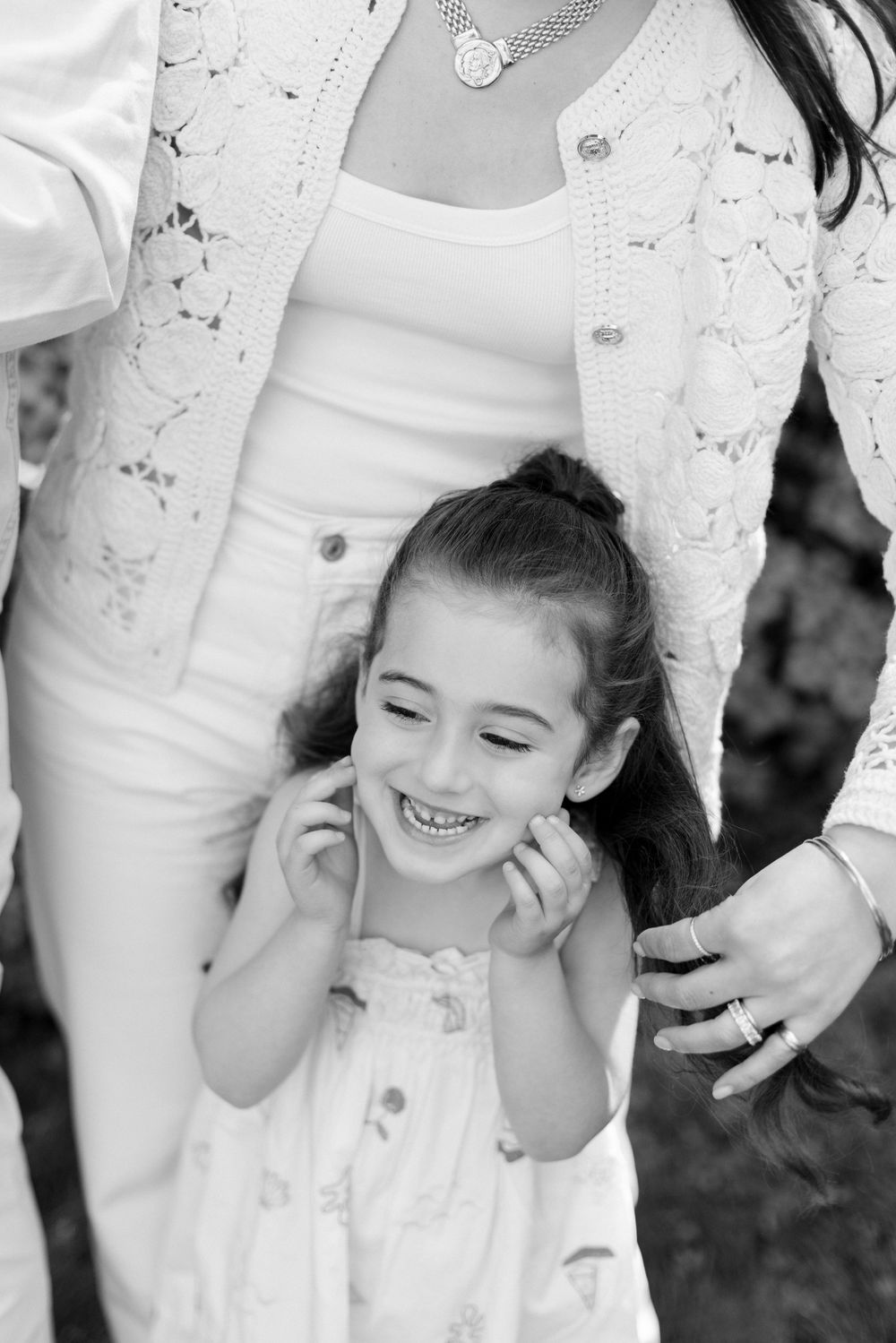 Black and white portrait of smiling child wearing white dress standing between adults in outdoor setting.