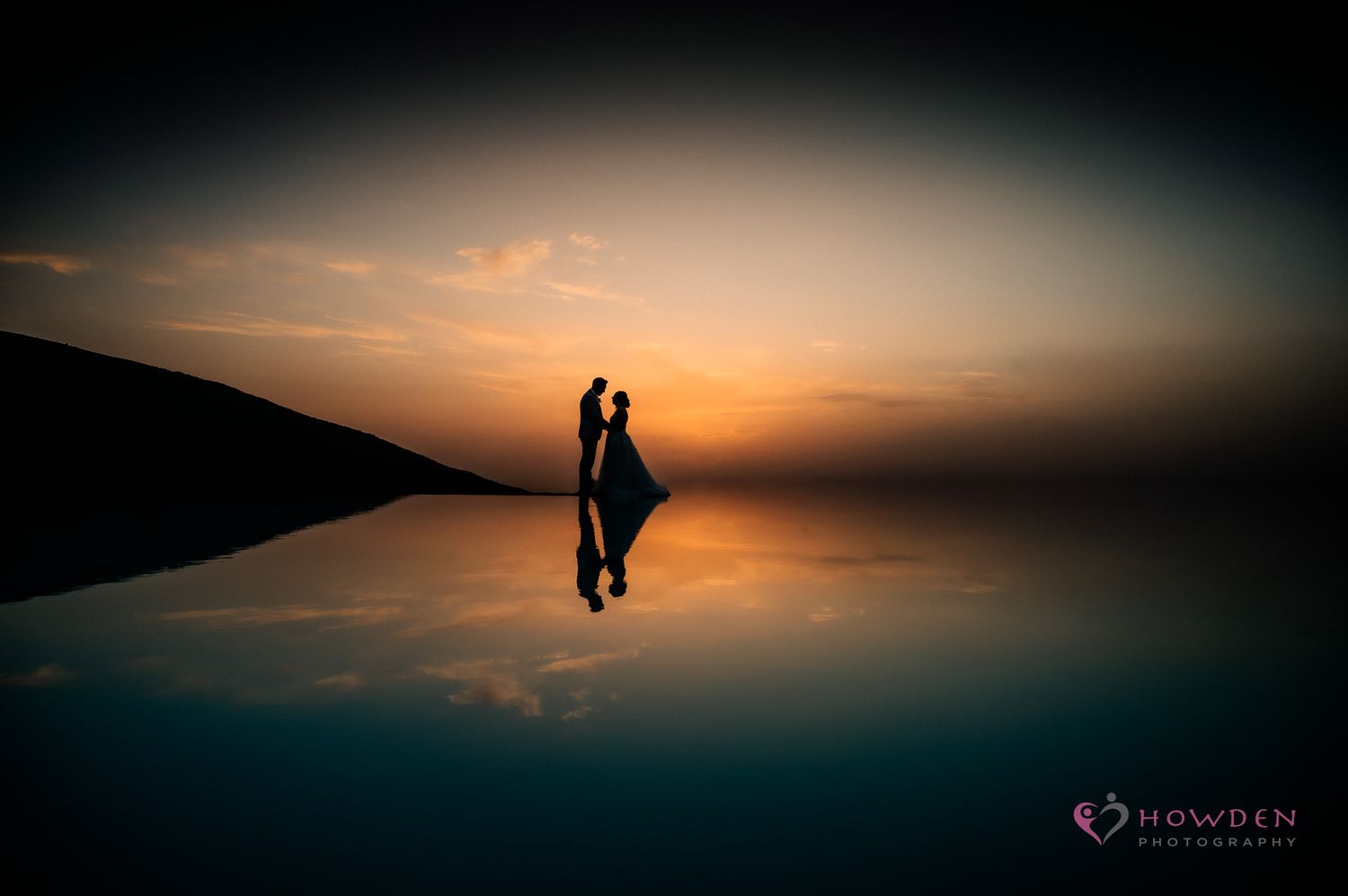 Silhouetted couple embraces against vibrant orange sunset sky on mountain ridge.