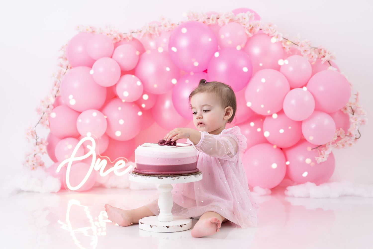 Baby in pink dress sits with cake against backdrop of pink balloons arranged in fan shape for first birthday photoshoot.
