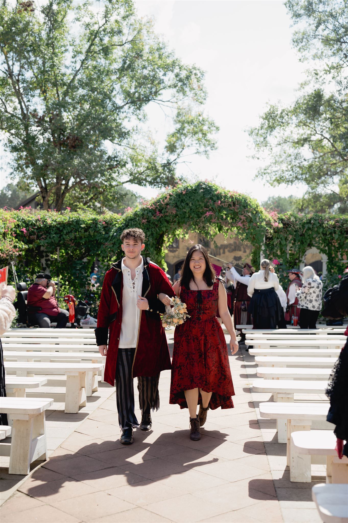 A couple in vintage-style red attire walks down an outdoor aisle lined with white chairs under a floral archway.