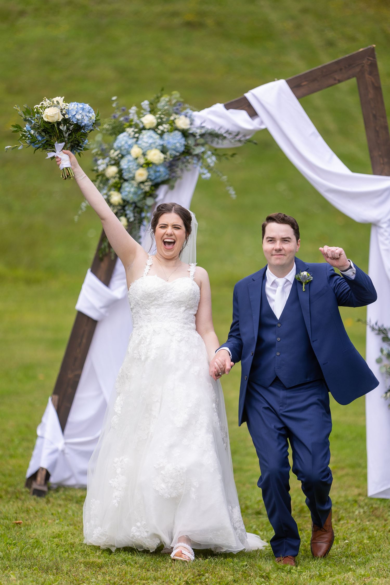 A joyful couple celebrates during their outdoor wedding ceremony under a wooden arch decorated with white fabric and flowers.