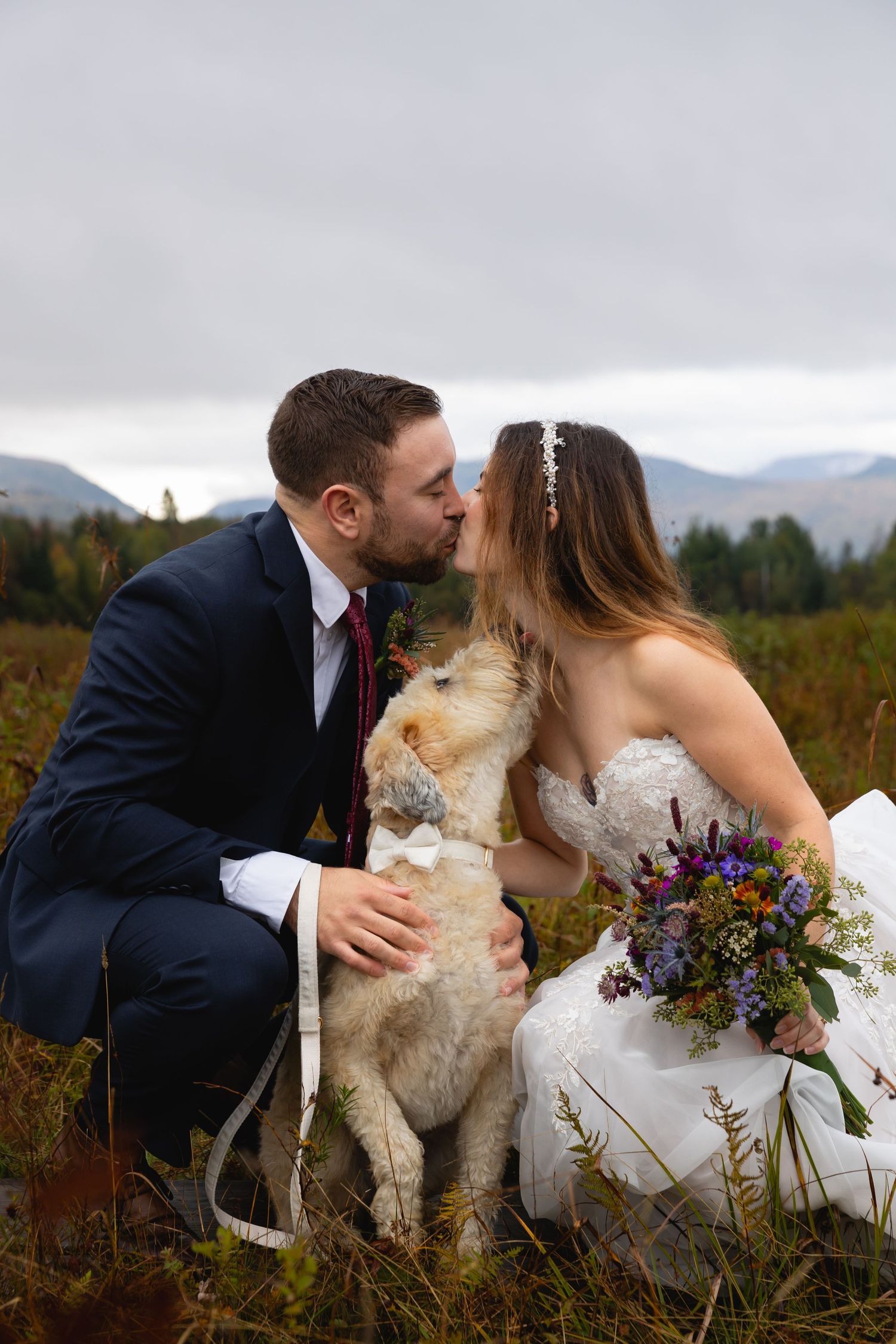 A bride and groom kiss outdoors in a mountain field while their happy dog sits between them with wildflower bouquet nearby.