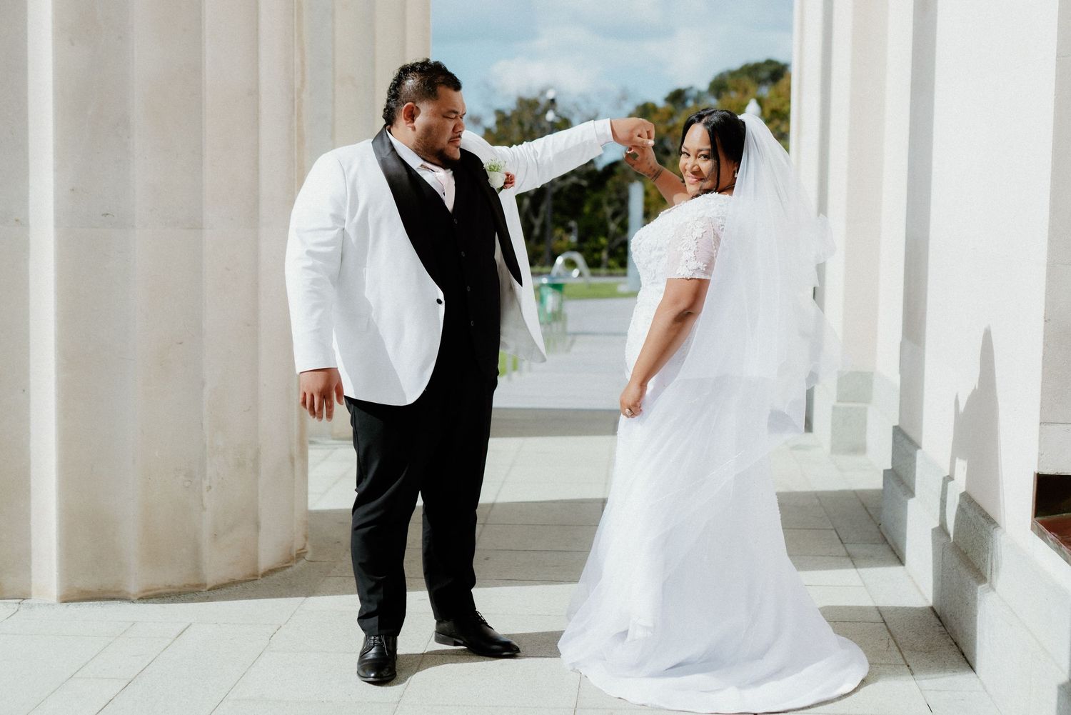 A couple shares a moment in a columned walkway on their wedding day, with the bride wearing a flowing white gown.