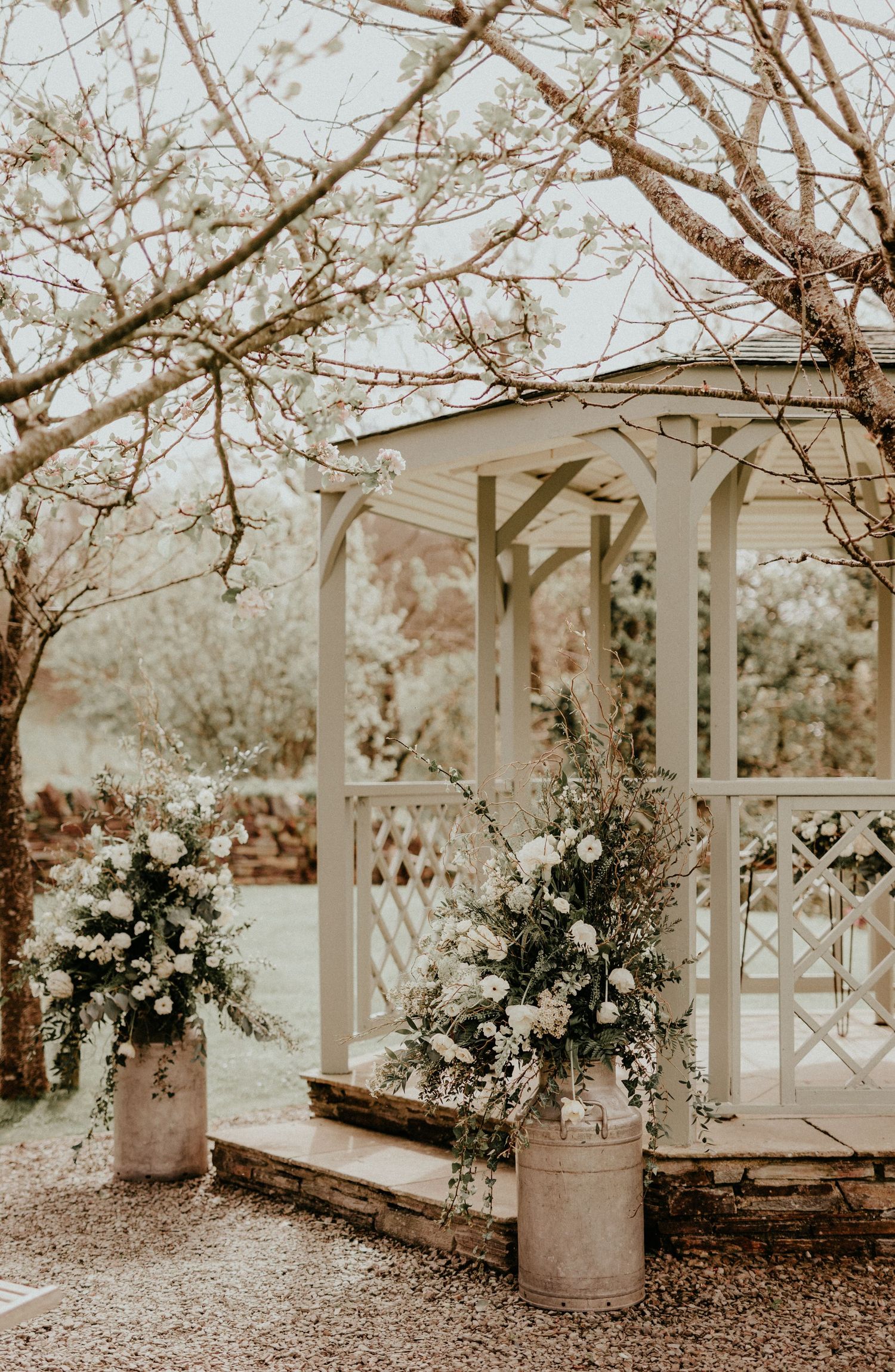 A vintage-style white gazebo decorated with floral arrangements in milk jugs under blooming spring tree branches.