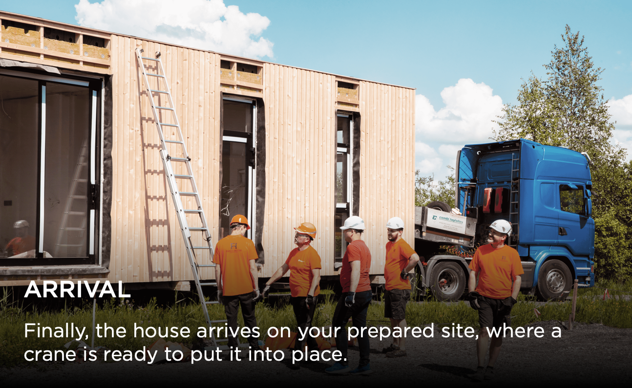 Construction workers use crane beams and forklifts to lift a modular house section inside a warehouse facility.