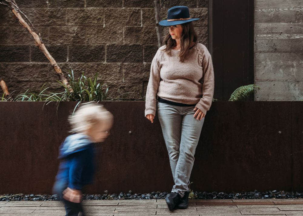 A person in a wide-brimmed hat and casual attire watches a blurred child running past a concrete wall with planters.