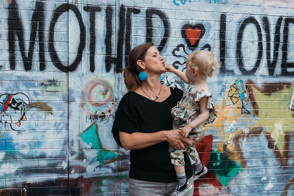 Someone in black holds child by graffiti wall with mother love text against colorful street art background.