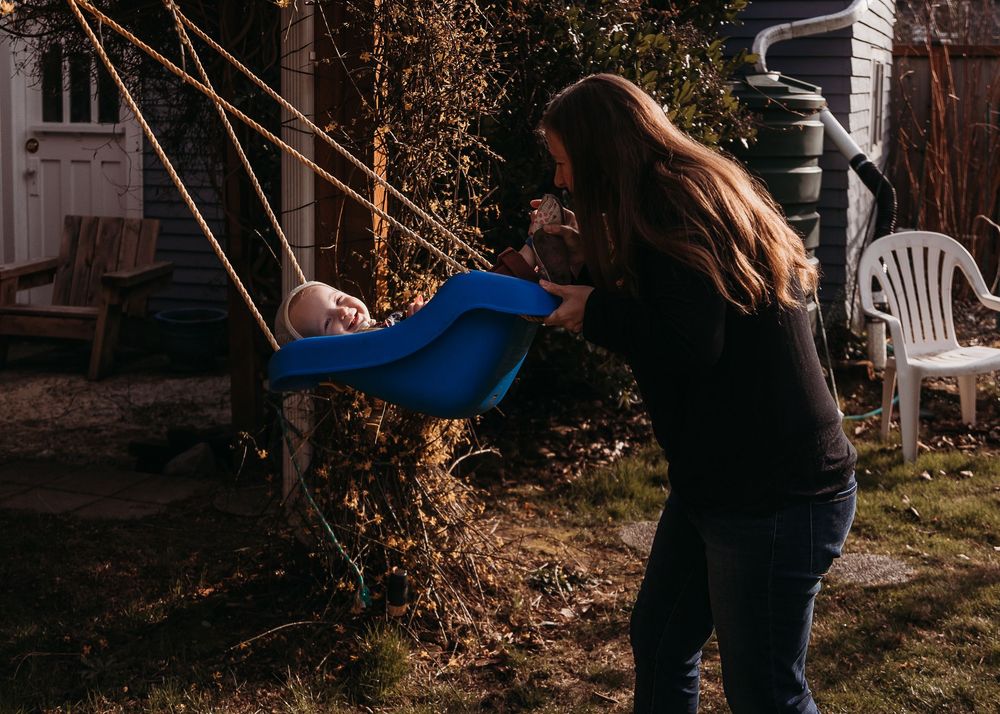 Someone pushes a child in a blue sweatshirt on a swing in a backyard during golden hour.