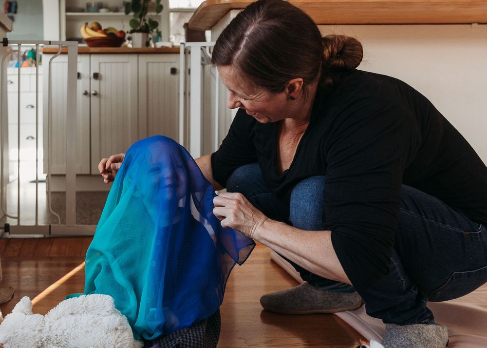A person wearing black clothing crouches down to sew something with blue fabric in a kitchen setting.