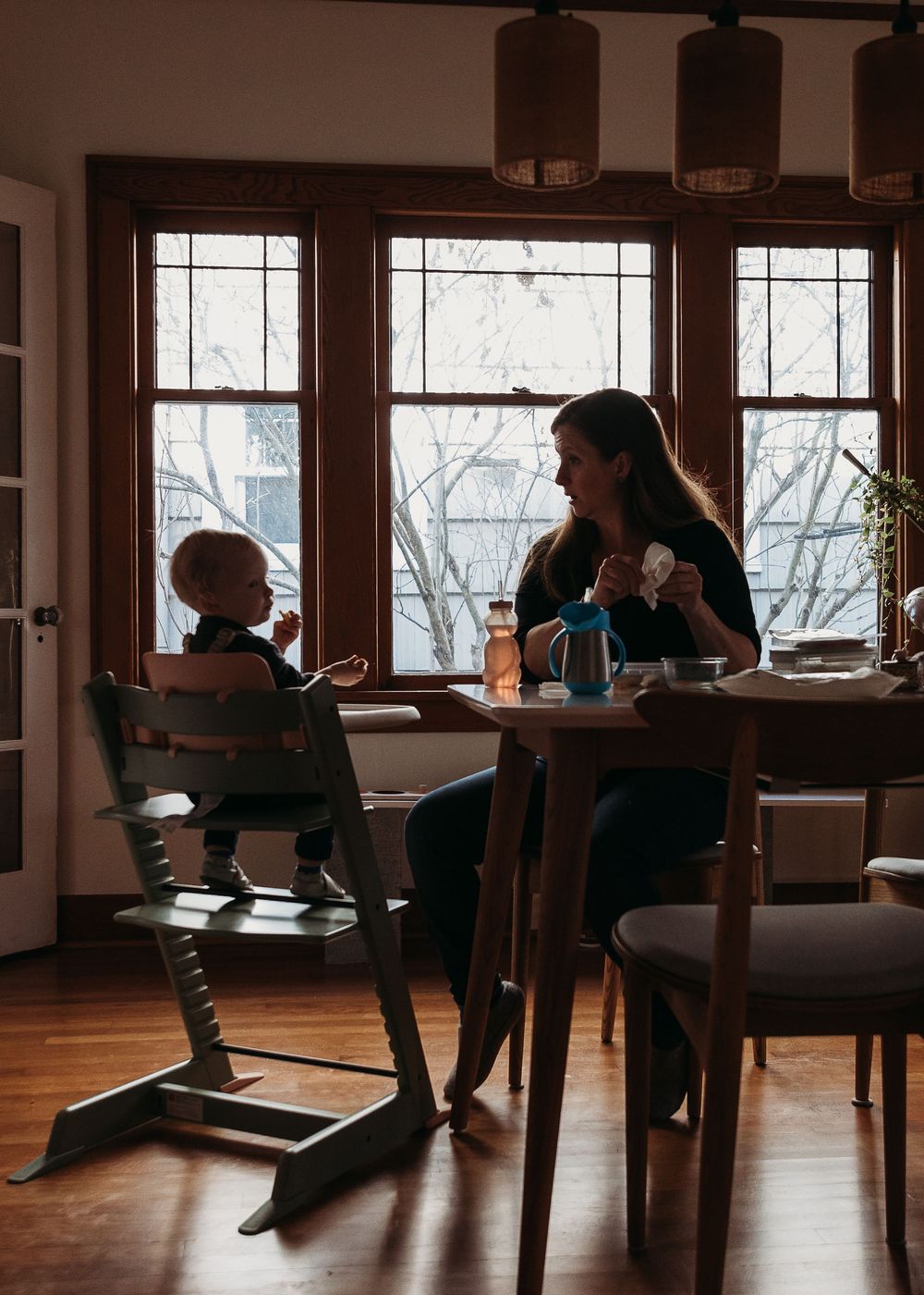 A silhouetted scene of a mealtime at home with warm lighting streaming through three windows in the background.