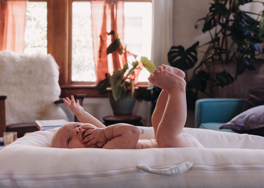 Baby lying on back playing with feet on white bedding in sunlit room with houseplants and window.