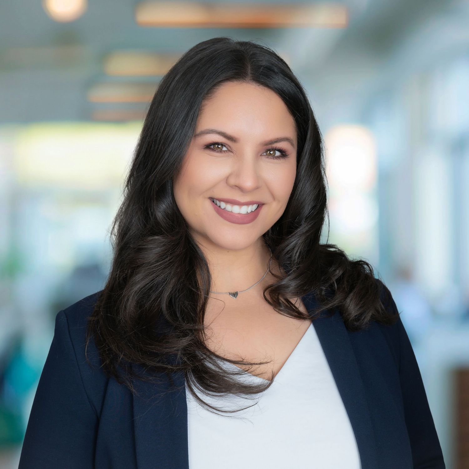 Professional portrait photo of someone with long dark hair wearing a navy blazer and white top in an office setting.