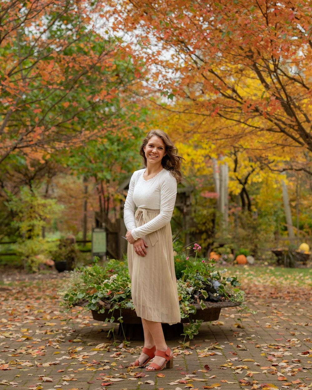 A person stands in an autumn garden wearing a cream sweater and beige pleated skirt amid colorful fall foliage.