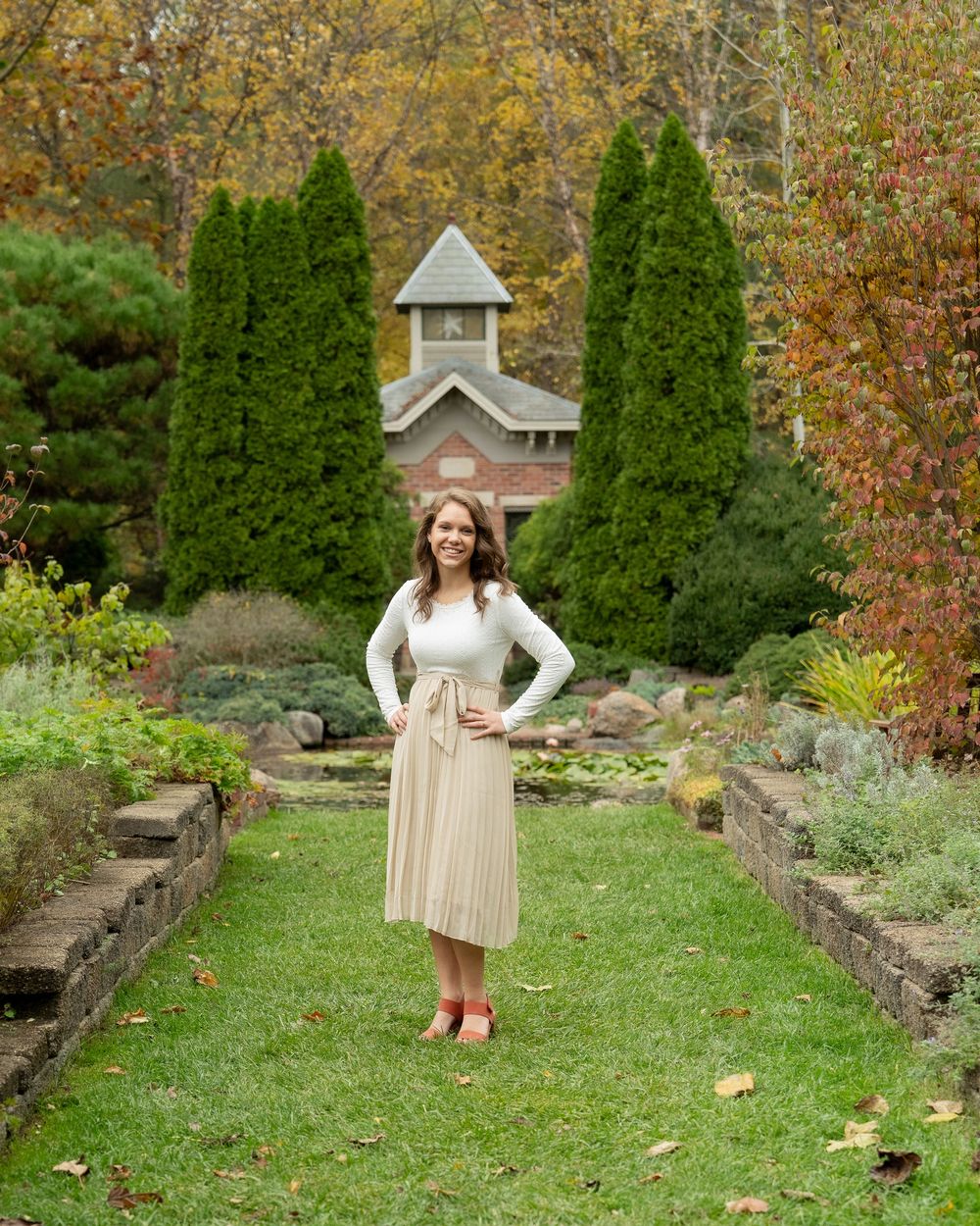 Someone poses in a cream-colored dress and cardigan on a garden path with evergreen trees and fall foliage background.