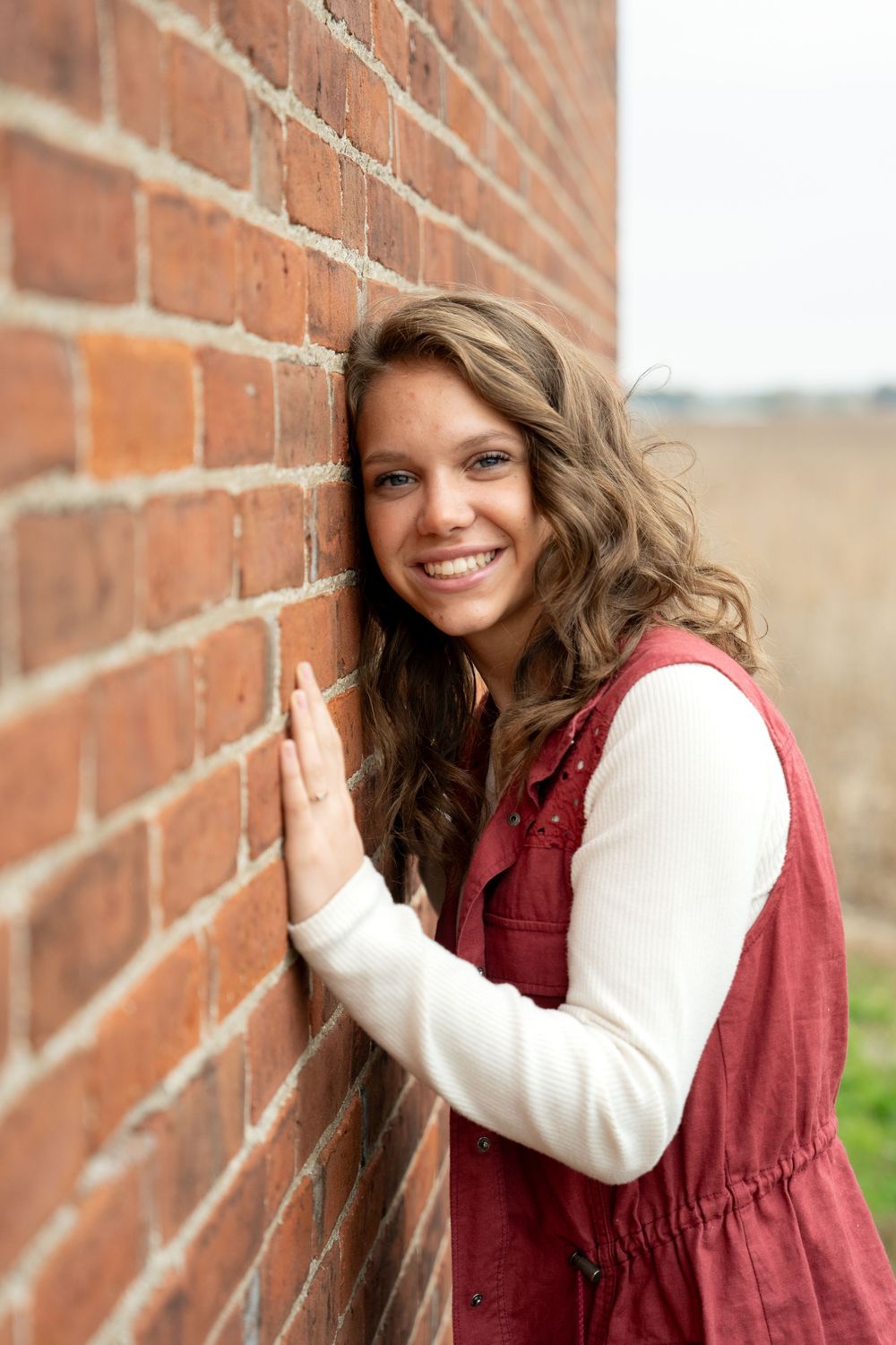 Portrait against brick wall showing wavy brown hair and warm smile wearing burgundy vest and cream sweater.