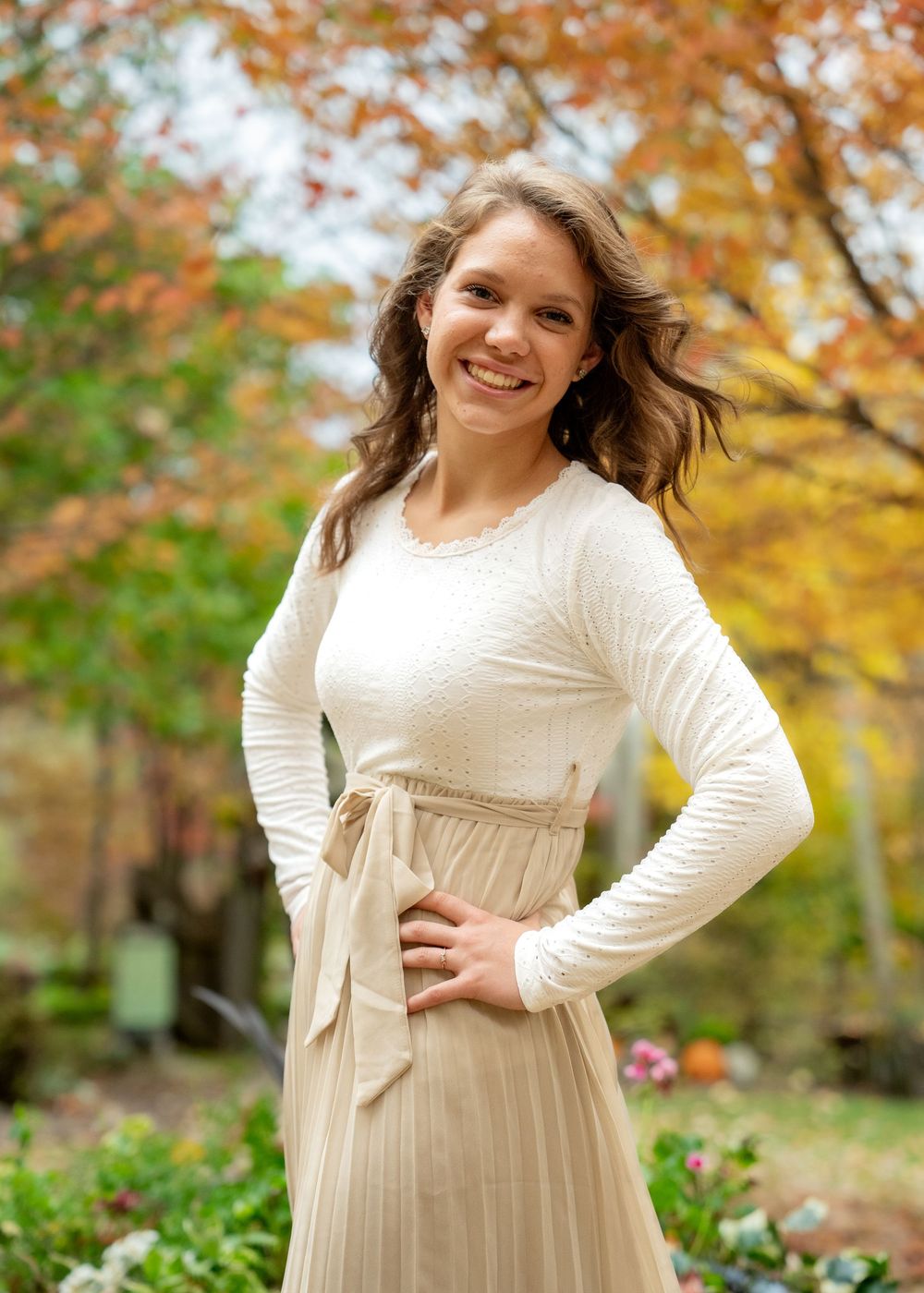 Person in a cream sweater and pleated skirt poses outdoors against autumn foliage with fall colors.