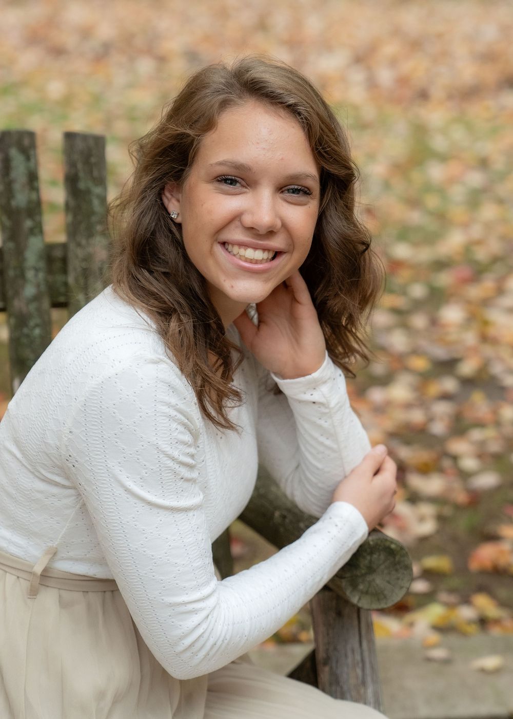 A person in a white sweater sits casually outdoors on a wooden bench surrounded by fallen autumn leaves.
