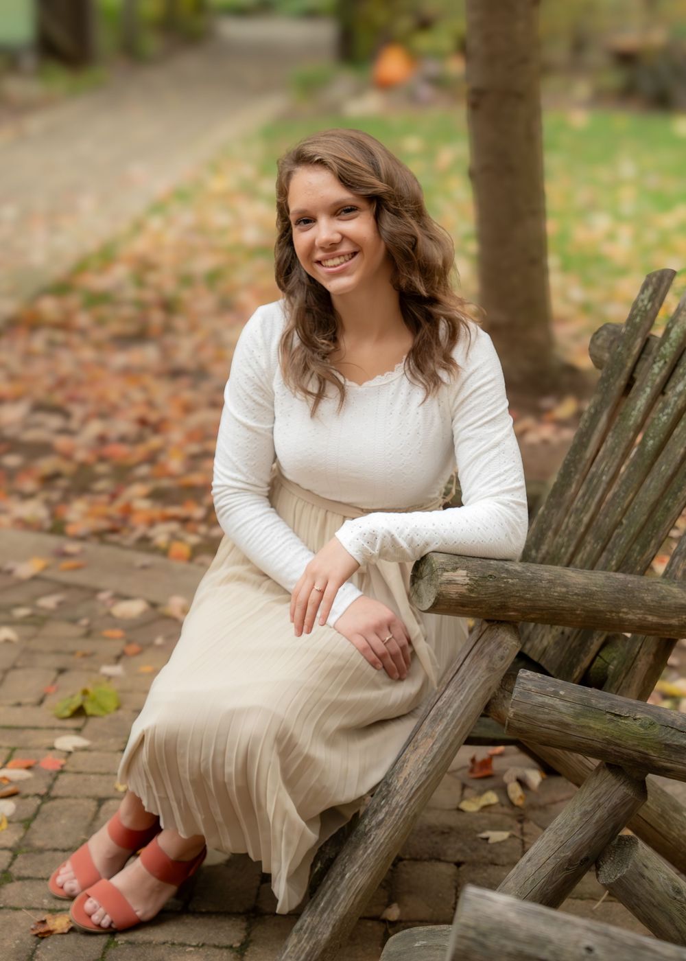 A person in a white long-sleeve top and beige dress sits on a rustic wooden fence surrounded by autumn leaves.