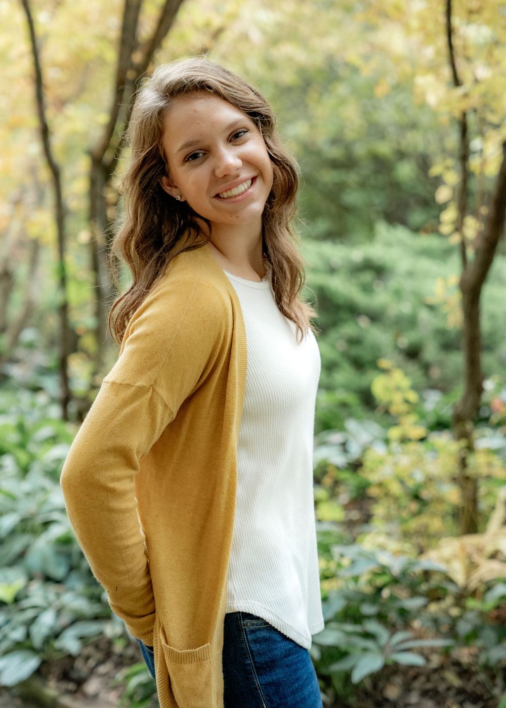 Person wearing mustard yellow cardigan and white top poses in an outdoor wooded setting with autumn foliage in background.