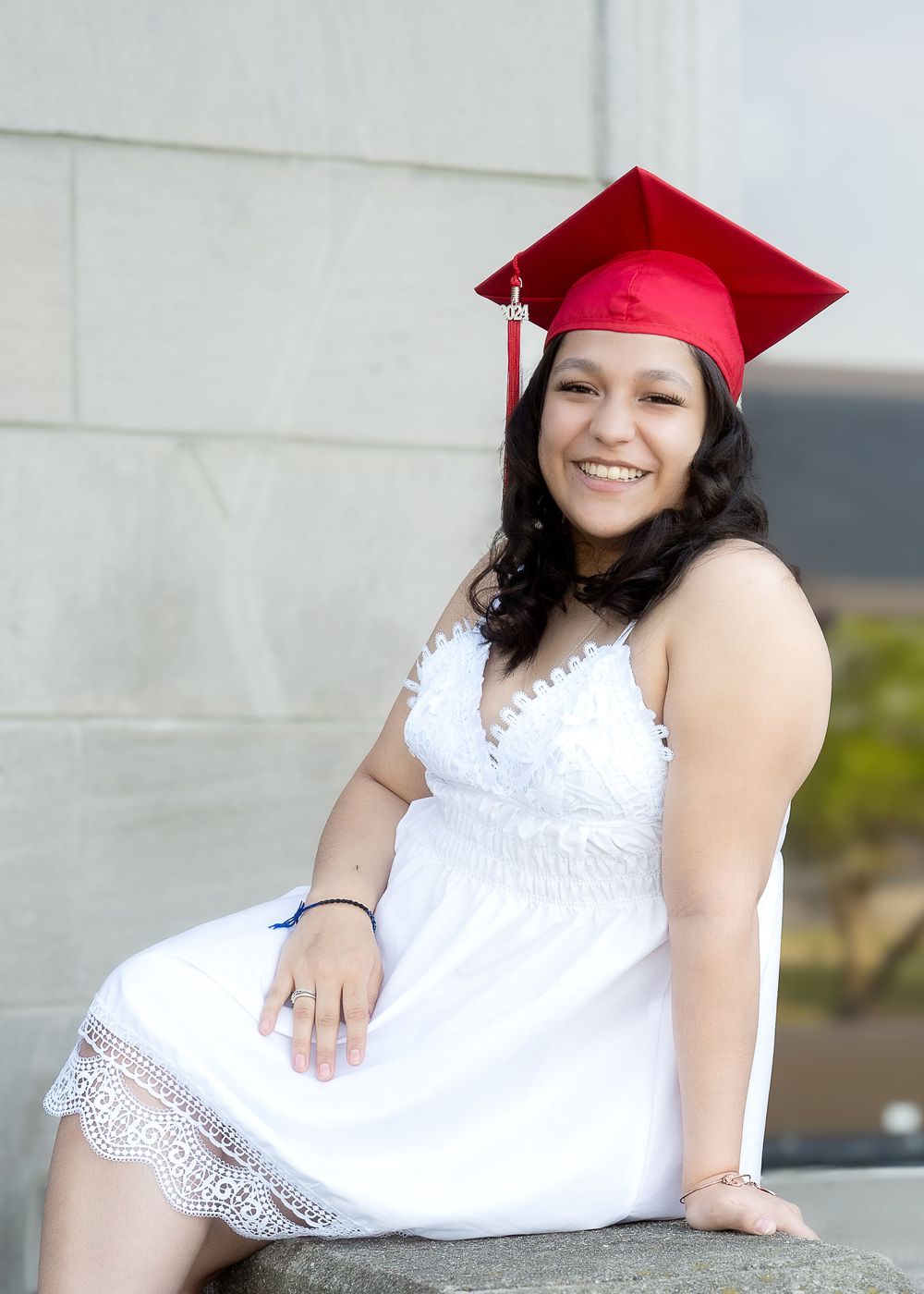 Graduate in red cap and white lace dress sits outdoors during commencement celebration.
