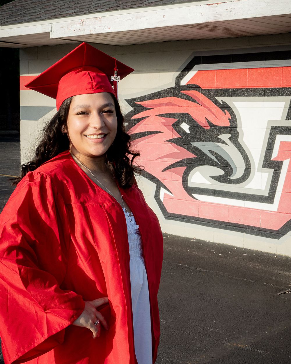 Graduate in red cap and gown poses in front of university sports logo on wall.