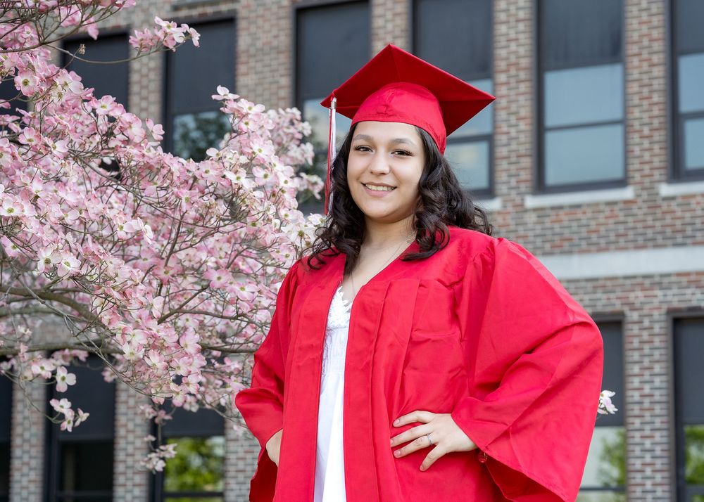 A graduate in red cap and gown poses by blooming cherry blossoms outside a brick building.