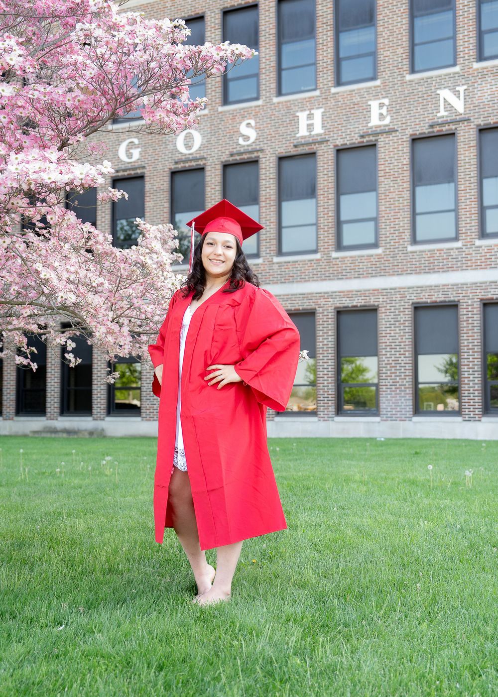 Graduate in red cap and gown poses on grass lawn in front of brick Goshen building with blooming cherry blossoms.
