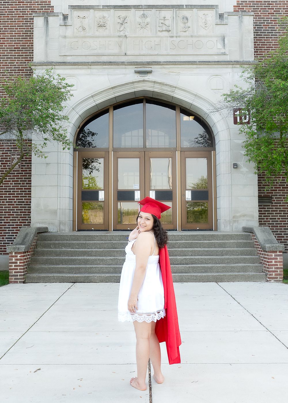 Graduate poses in red cap and white dress in front of stone archway entrance to brick academic building.