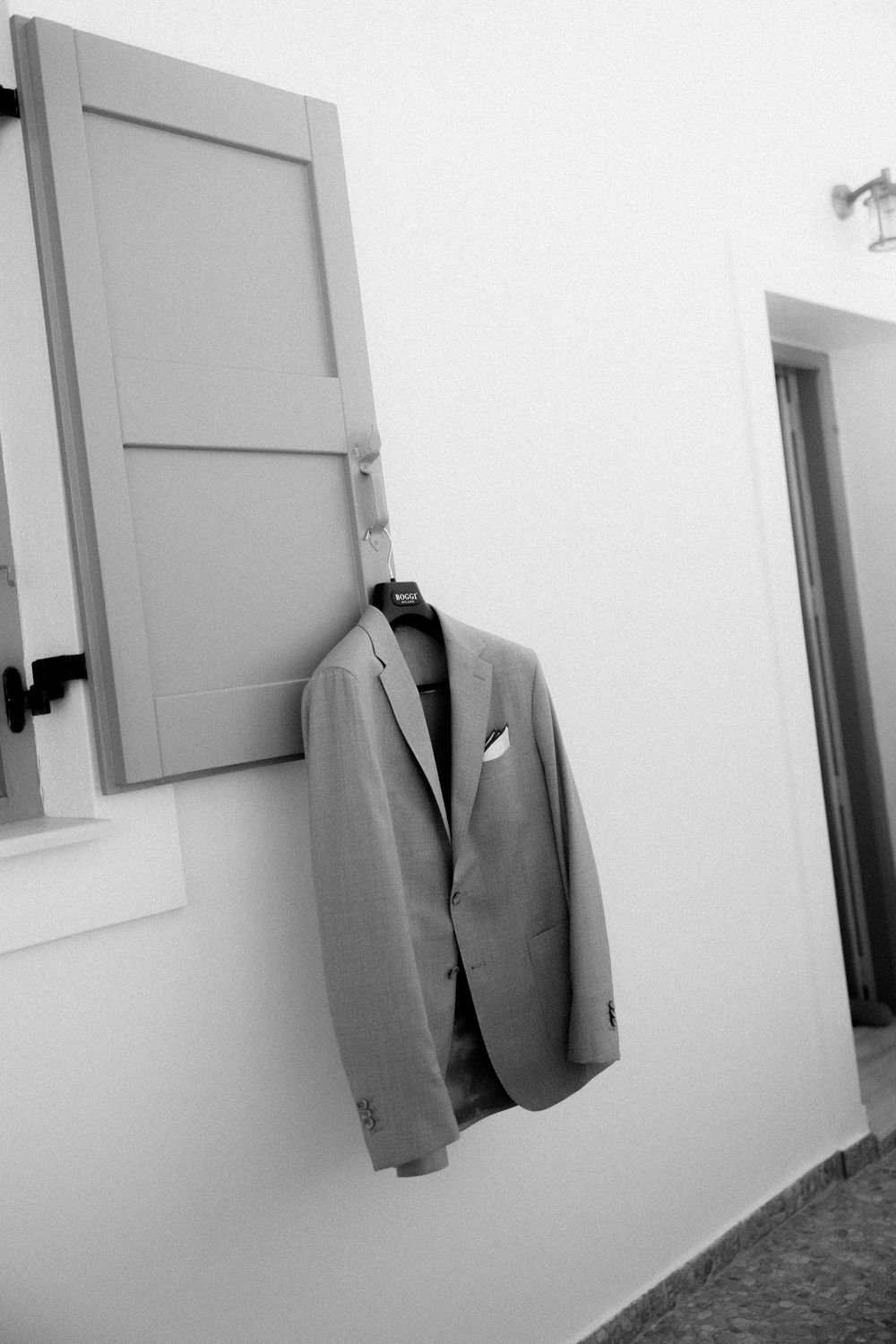 Black and white photo of a grey suit jacket hanging on a white wall. Astypalaia wedding photographer Greece