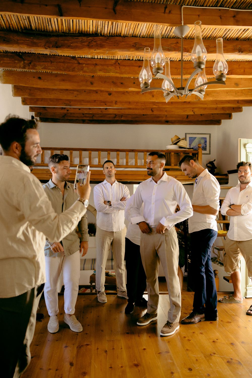 Group of people in white and beige attire gathered in a rustic room with wooden beams and chandelier. Astypalaia wedding photographer Greece