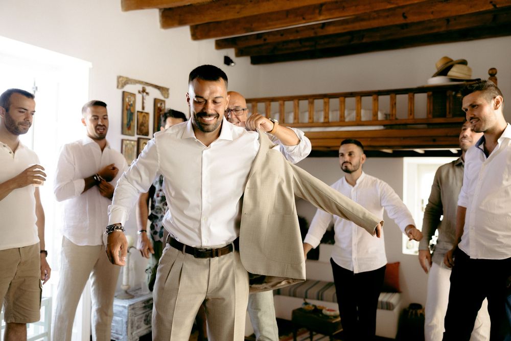 People helping someone put on a light colored suit jacket in a rustic indoor setting. Astypalaia wedding photographer Greece