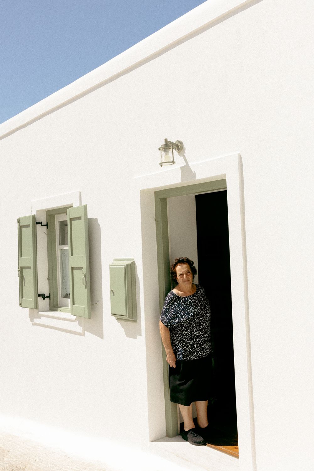 Person standing in doorway of white Mediterranean-style building with green shutters. Astypalaia wedding photographer Greece