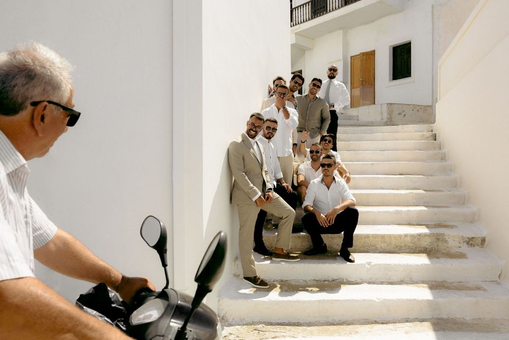 People in formal attire posing on white steps while someone on scooter passes by. Astypalaia wedding photographer Greece