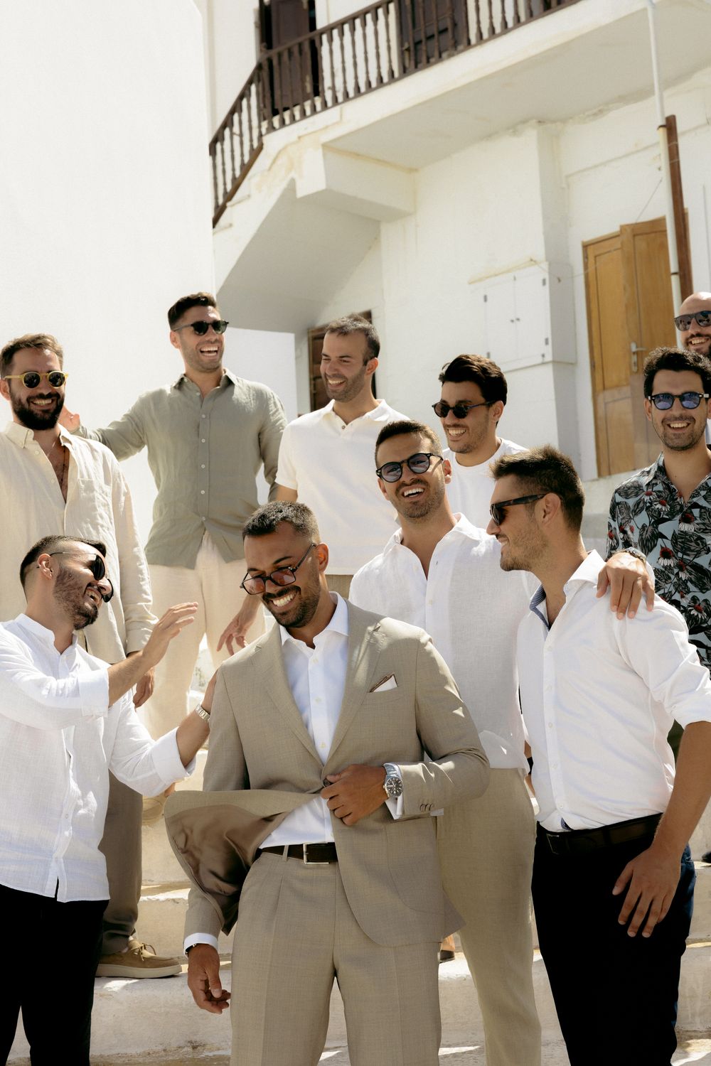 Groomsmen and groom in light colored formal wear laughing and posing together against white building. Astypalaia wedding photographer Greece