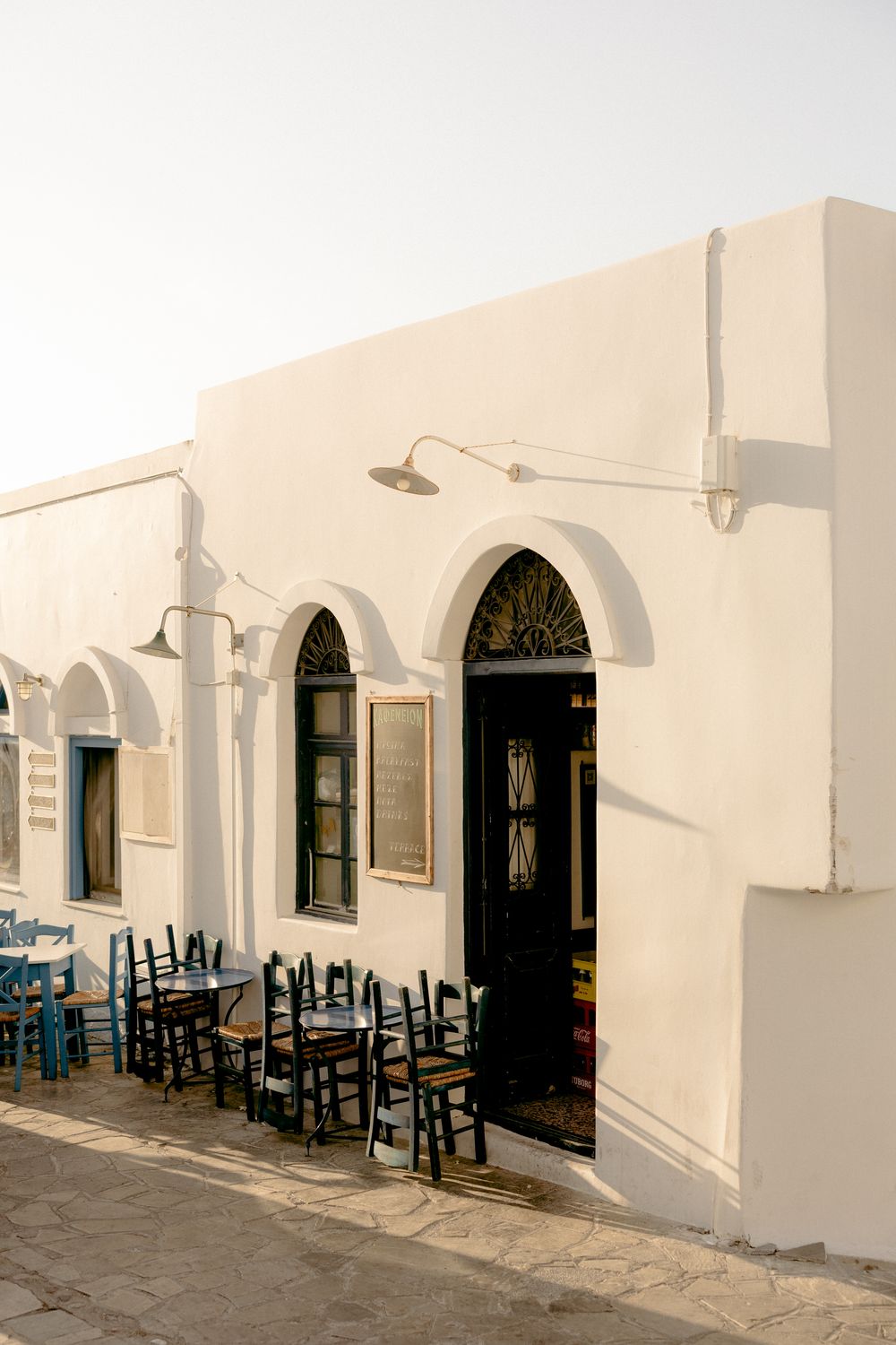 White Mediterranean cafe exterior with blue chairs and arched doorways.