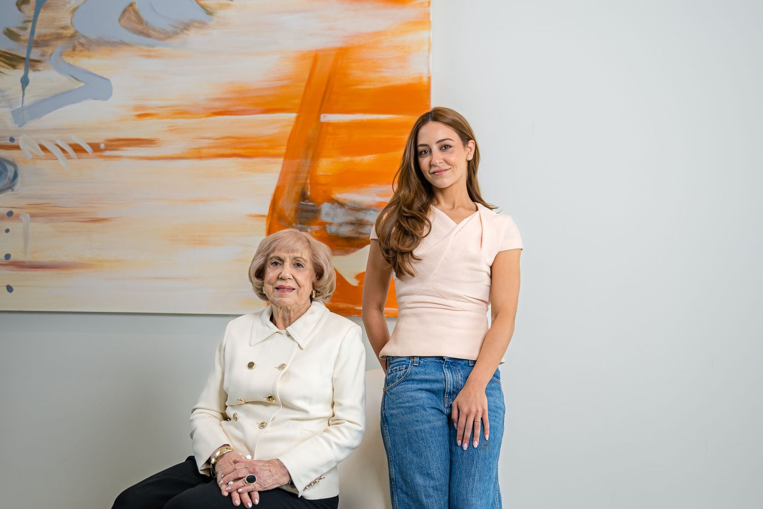 Two people pose against a wall with an abstract orange painting, one seated in white and one standing in casual attire.