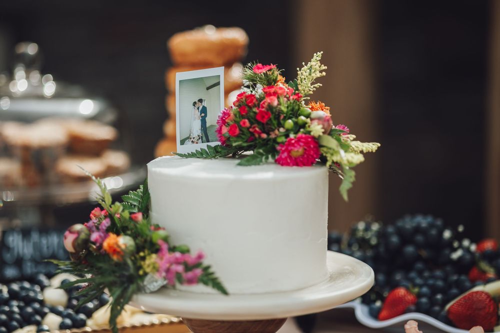 White wedding cake decorated with fresh colorful flowers and a small topper sits on an elegant table setting.