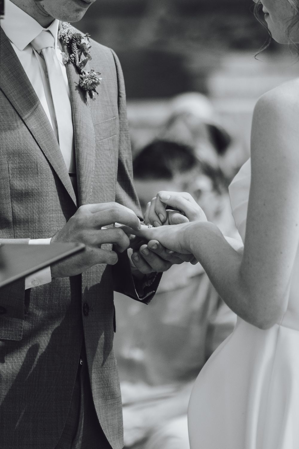 Black and white wedding ceremony photo of couple exchanging rings during intimate outdoor ceremony.
