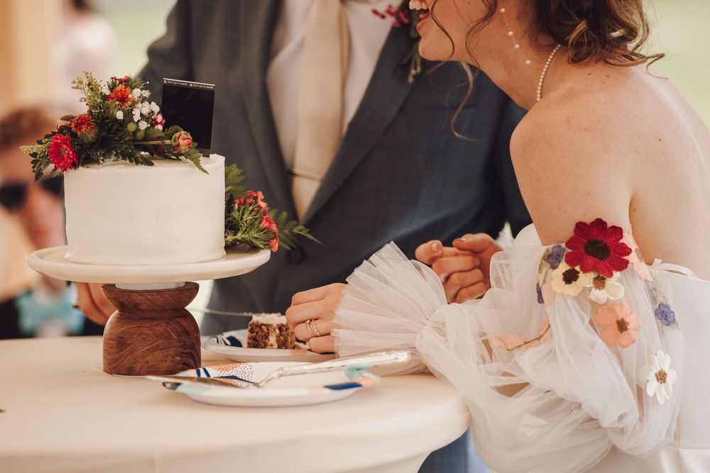 A couple cuts their white wedding cake decorated with red flowers on a wooden stand at their reception.