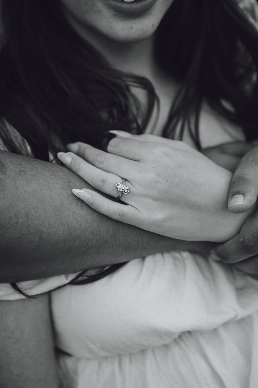 Black and white close-up of hands showing an engagement ring against white fabric.