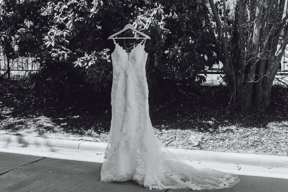 An elegant white lace wedding dress with a fitted silhouette hangs outdoors in a black and white photograph.