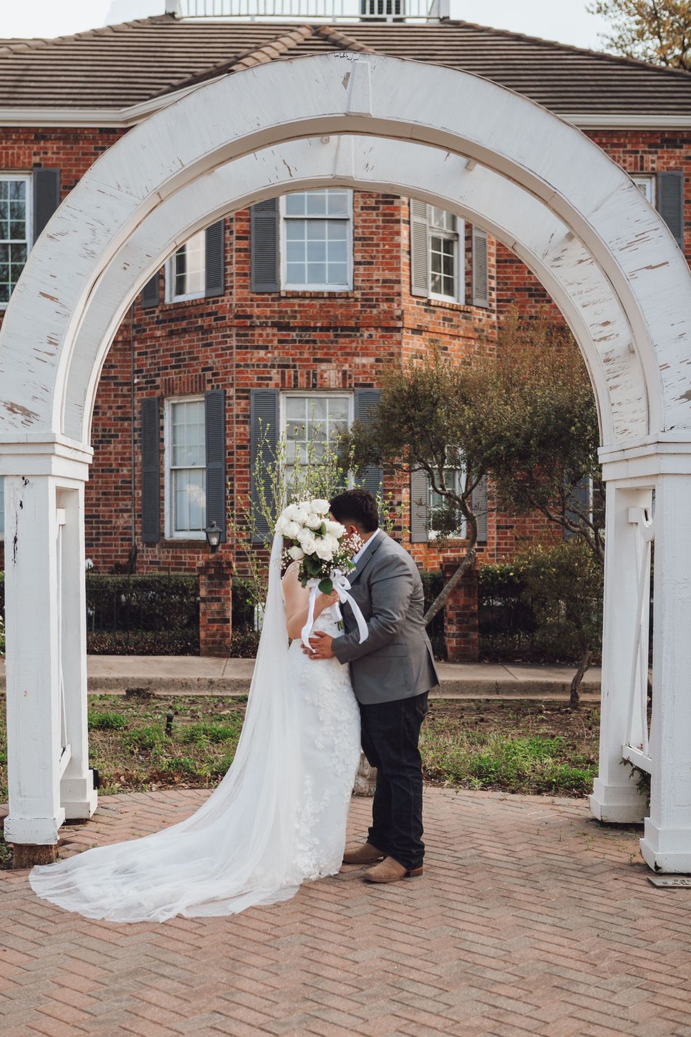Wedding photos captured through white archways on brick paver walkway with colonial brick building in background.