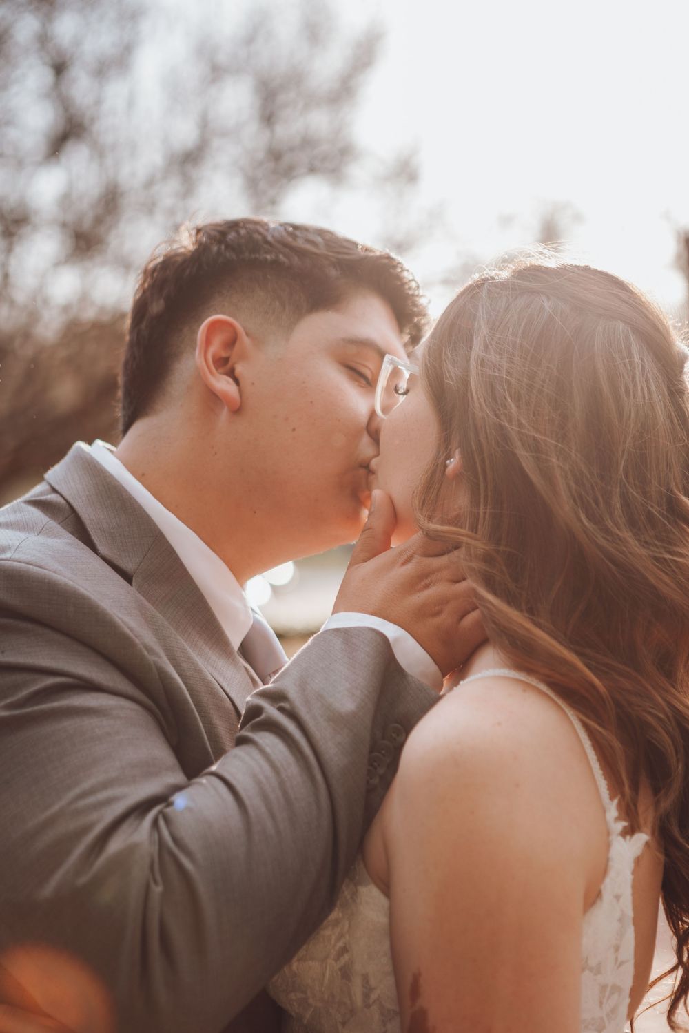 A tender romantic moment between newlyweds sharing a kiss against a warm sunlit backdrop in formal wedding attire.