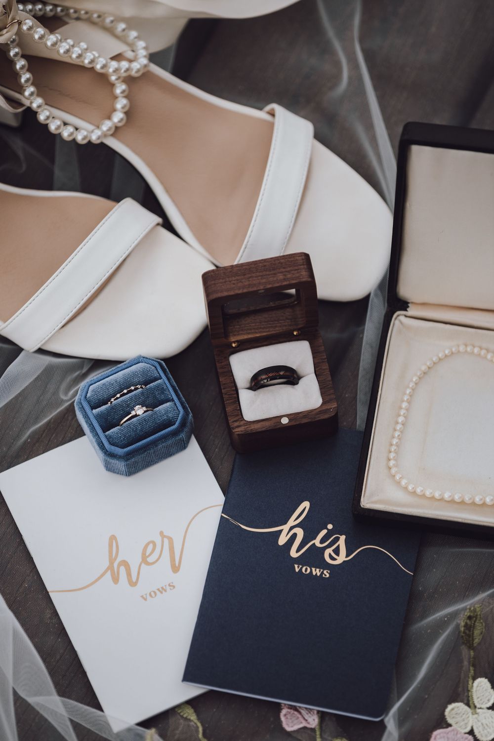 Wedding details flatlay featuring white bridal shoes, his and her vow books, ring boxes, and pearl jewelry on gray fabric.