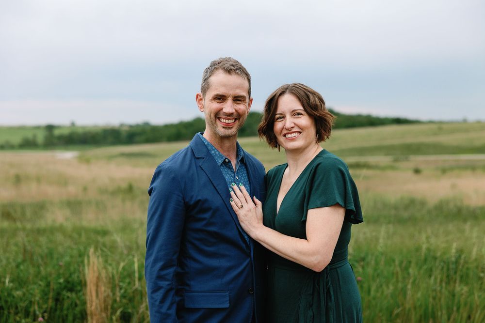 A man and woman stand side-by-side smiling in the middle of the Neal Smith priaire, her left hand on his chest showing her engagement ring.