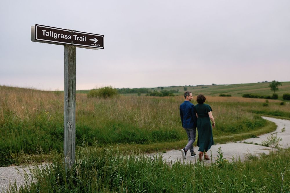 A couple walks down a paved path through the Iowa prairie at Neal Smith Wildlife Refuge, a trail sign reading "Tallgrass Trail" in front