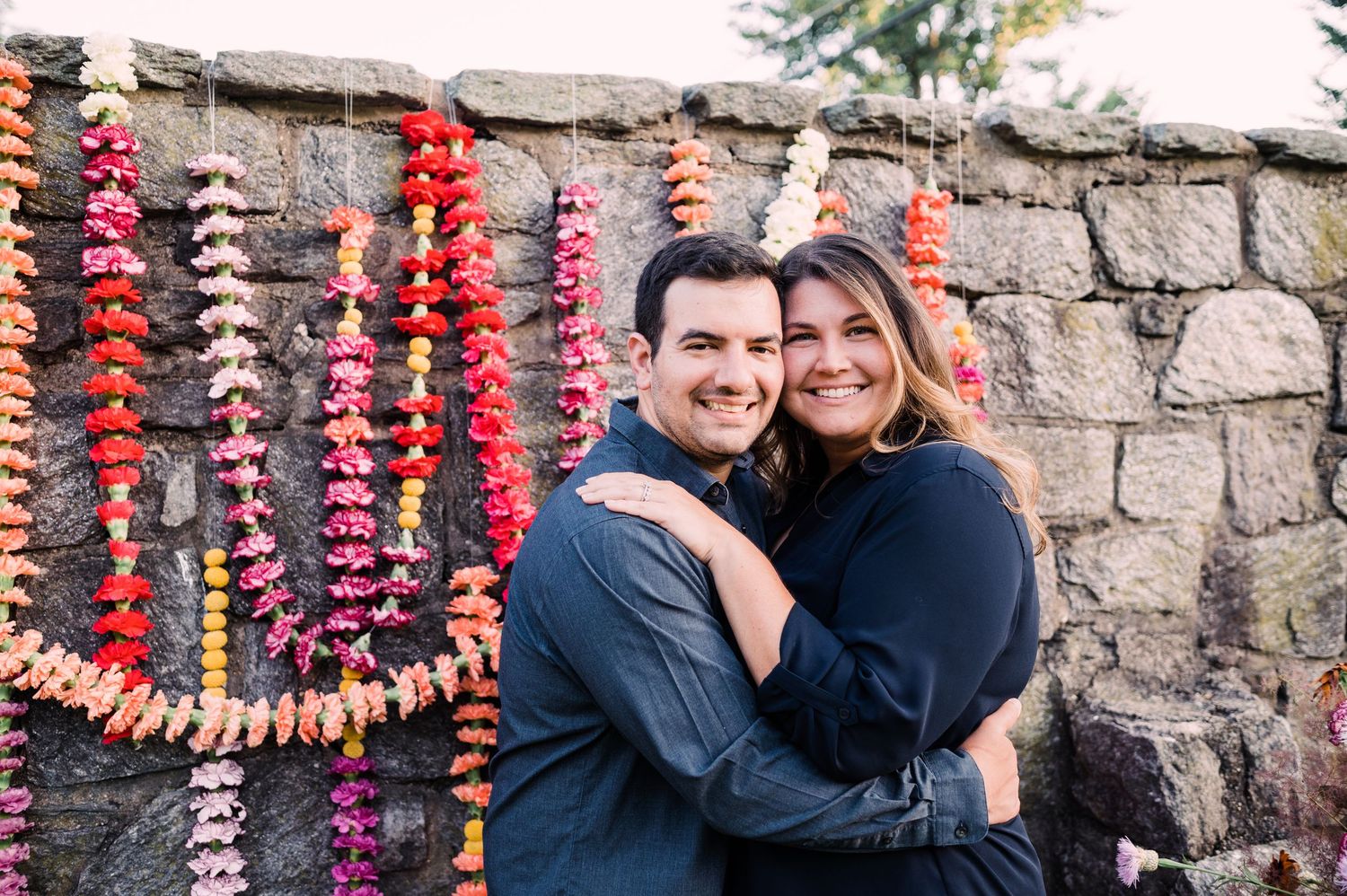 A couple embraces in front of a stone wall decorated with colorful hanging paper flower garlands.